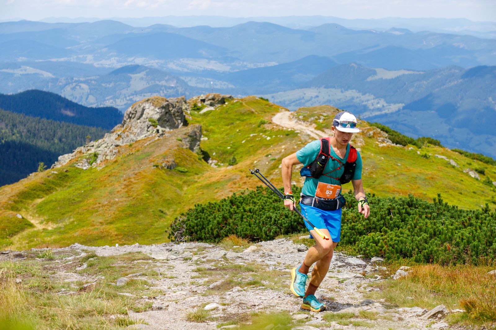 Trail runner wearing a hydration vest and carrying folded poles on a rocky mountain path.