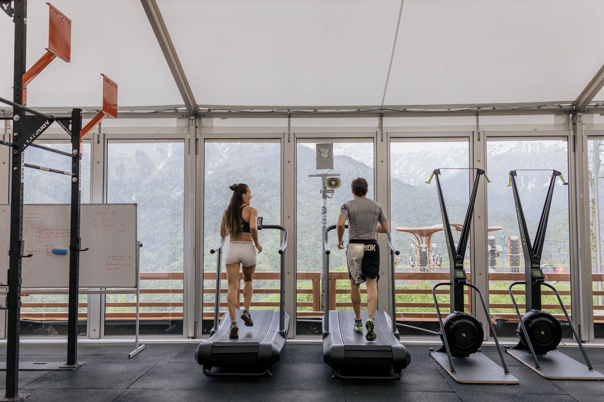Two people using treadmills indoors beside large windows.